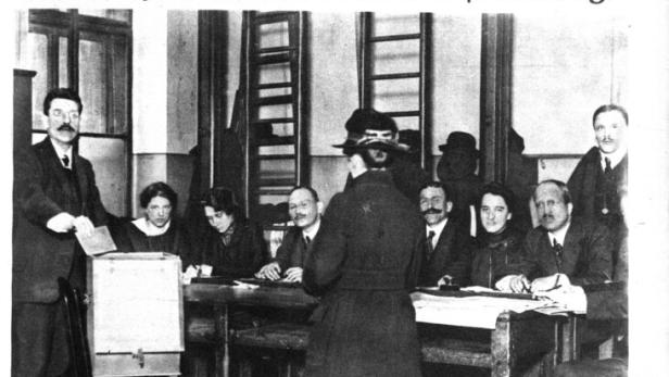 Mehrere Menschen sitzen an Tischen, während eine Frau in historischer Kleidung an einer Wahlurne steht und wählt.