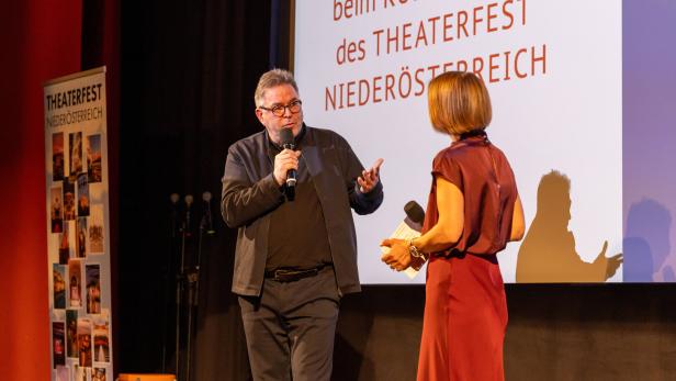 Ein Mann und eine Frau sprechen auf einer Bühne mit Mikrofonen vor einer Leinwand mit Text zum Theaterfest Niederösterreich.