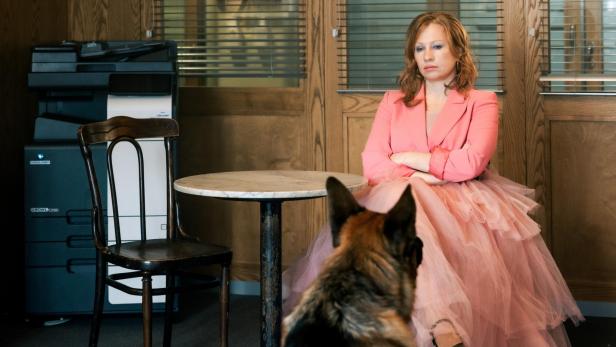 Eine Frau im rosa Tüllkleid sitzt mit verschränkten Armen an einem Tisch, ein Schäferhund sitzt ihr gegenüber.