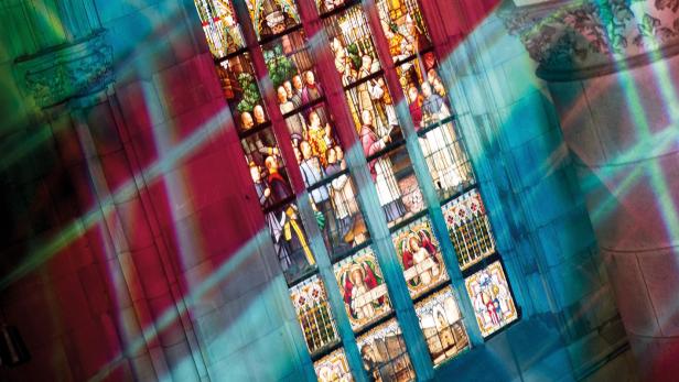 Buntes Licht fällt durch ein großes Kirchenfenster mit detailreichen Glasmalereien und Figuren auf steinerne Wände.