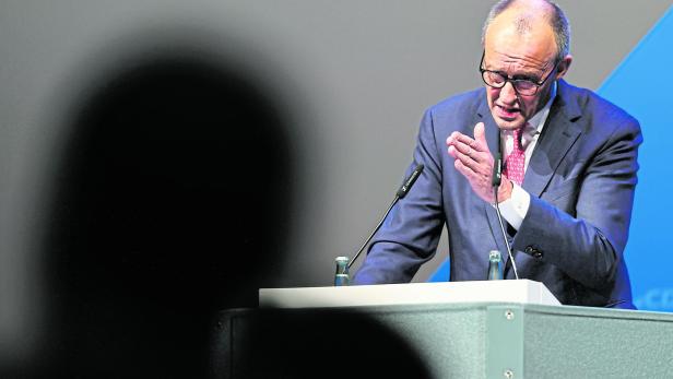 Ein Mann im Anzug spricht gestikulierend an einem Rednerpult mit Mikrofon und Wasserflaschen.