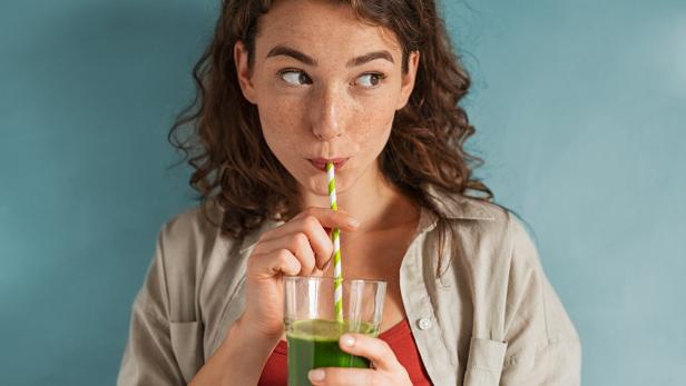 Frau mit lockigen Haaren trinkt mit einem gestreiften Strohhalm ein grünes Getränk aus einem Glas vor blauem Hintergrund.