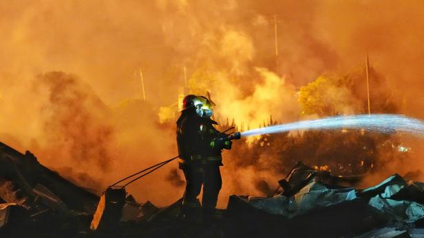 Zwei Feuerwehrleute löschen mit einem Wasserschlauch ein großes Feuer inmitten dichter Rauchwolken.