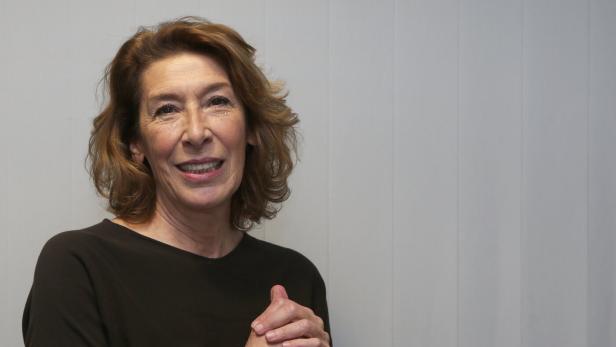 Eine lächelnde Adele Neuhauser mit braunen Haaren und dunklem Oberteil hält die Hände vor einem hellen Hintergrund verschränkt.