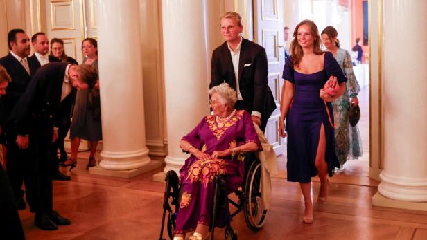 Prinzessin Astrid im Rollstuhl wird von einem jungen Mann begleitet, gefolgt von weiteren Personen.