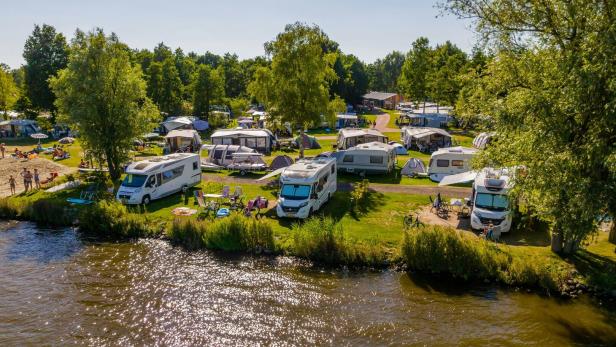 Ein Campingplatz am Wasser mit Wohnwagen, Zelten und Badegästen unter blauem Himmel.