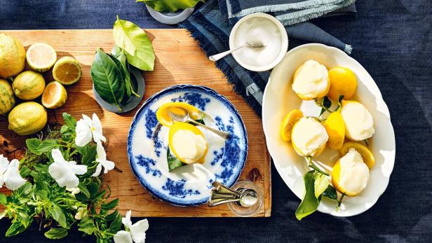 Zitronensorbet wird in ausgehöhlten Zitronenhälften serviert, daneben frische Zitronen, Blätter und weiße Blumen.