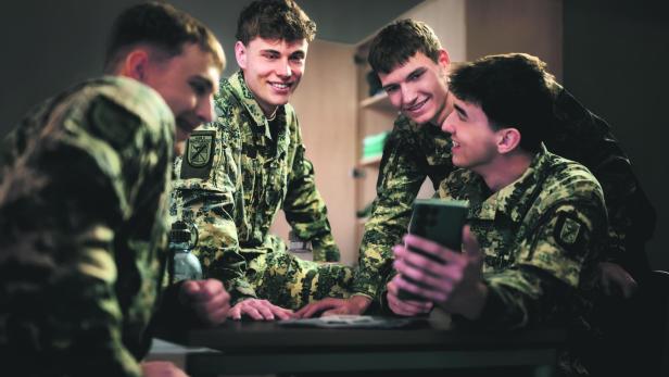 Eine Gruppe junger Männer in Bundesheer-Uniform sitzen, schauen  auf einen Handybildschirm und lachen.