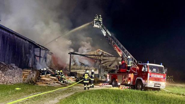 Feuerwehrleute löschen nachts mit einem Löschfahrzeug und einer Drehleiter einen brennenden Holzstapel an einem Gebäude.