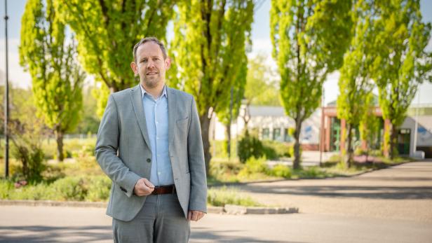 Ein Mann im grauen Anzug steht auf einer sonnigen Straße vor grünen Bäumen und einem Gebäude im Hintergrund.