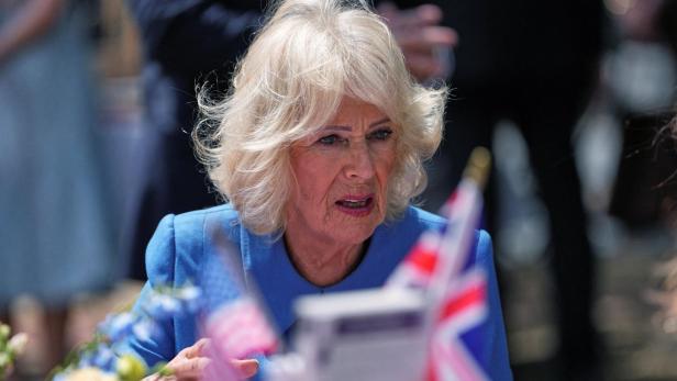 Königin Camilla, in einem blauen Blazer, spricht vor britischen Flaggen.