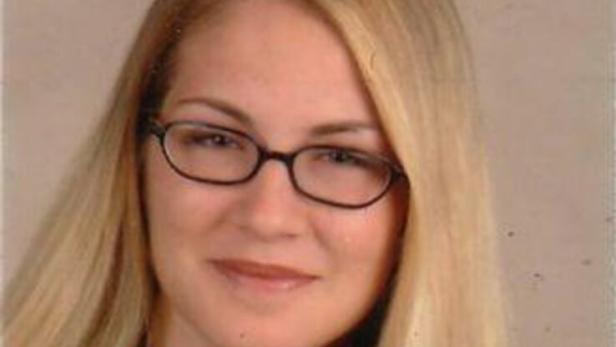 Junge Frau mit langen blonden Haaren und Brille, trägt ein dunkles Oberteil und lächelt leicht vor neutralem Hintergrund.