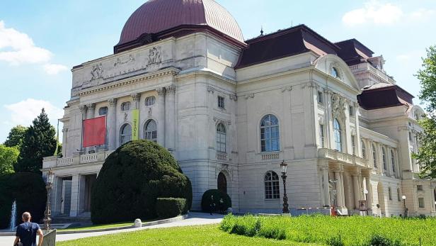 Ein großes, historisches Gebäude mit Kuppeldach und verzierten Fassaden steht inmitten einer grünen Parkanlage.