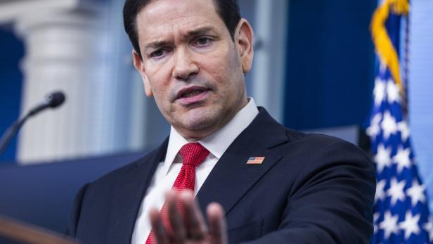 Marco Rubio spricht vor einer US-Flagge und trägt einen dunklen Anzug mit roter Krawatte.