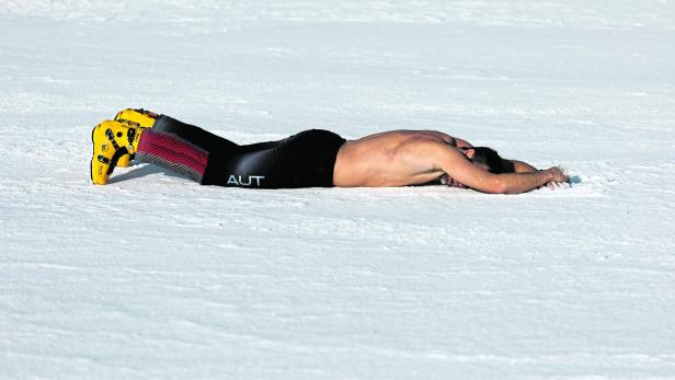 Ein Mann liegt oberkörperfrei ausgestreckt mit gelben Skischuhen auf dem Schnee.
