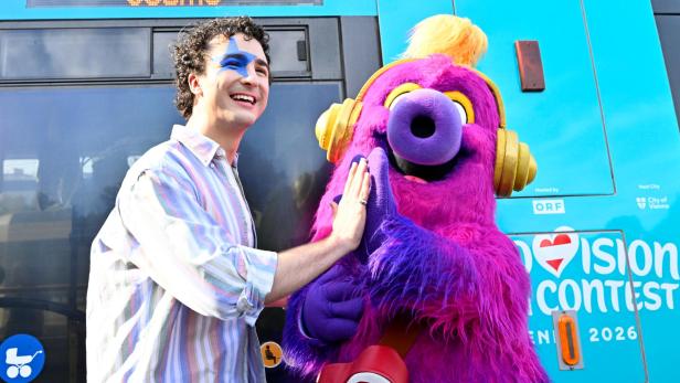 Ein lachender Mensch mit blauem Gesichtsmal und ein großes, lila Plüschmaskottchen geben sich ein High-Five vor einem bunten Bus.