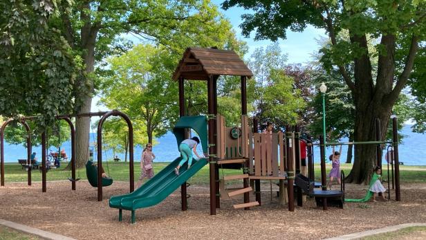 Kinder spielen auf einem Spielplatz unter großen, schattenspendenden Bäumen.