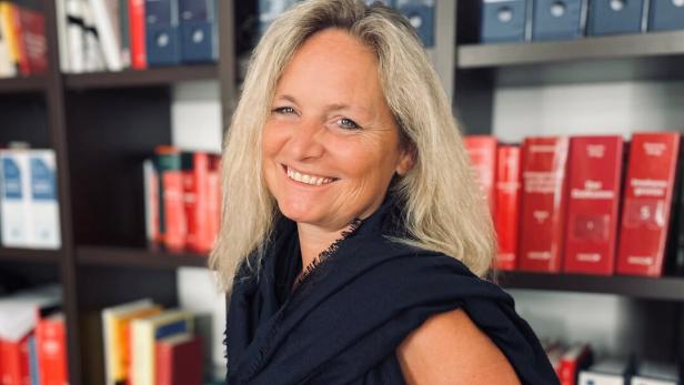 Eine lächelnde Frau mit blonden Haaren steht vor einem Bücherregal.