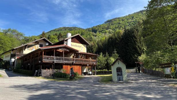 Ein Gasthaus mit Holzveranda steht vor bewaldeten Hügeln, daneben eine kleine Kapelle und umgeben von grünen Bäumen.