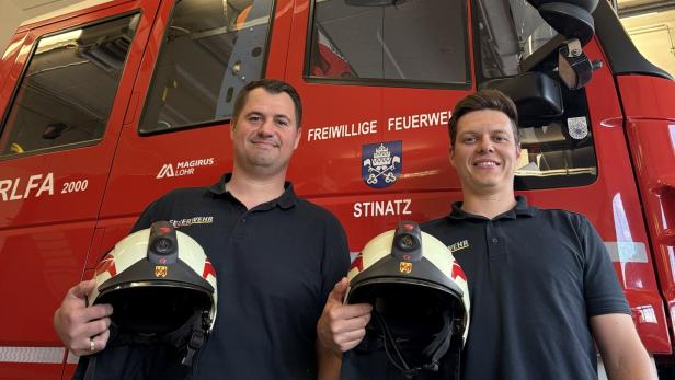 Zwei Feuerwehrmänner stehen vor einem roten Feuerwehrfahrzeug und halten jeweils einen Feuerwehrhelm in der Hand.