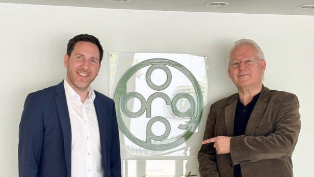 Zwei Männer stehen nebeneinander vor einem Wandlogo, einer zeigt auf das Logo, beide lächeln.