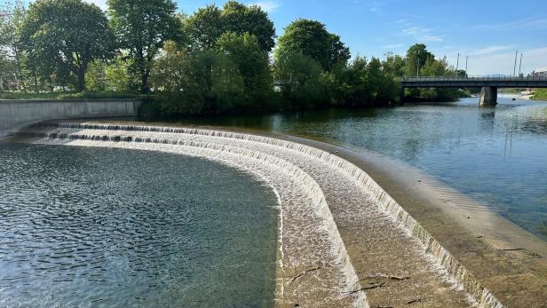 Wasser fließt in mehreren Stufen über ein Wehr, im Hintergrund Bäume und eine Brücke unter blauem Himmel.