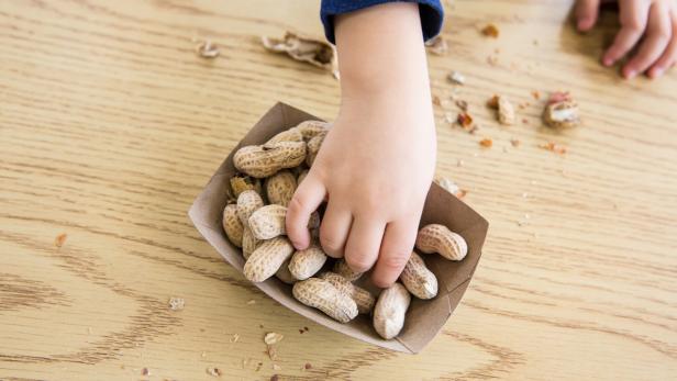 Eine Kinderhand greift nach Erdnüssen in einer Pappschale auf einem Holztisch mit Erdnussschalenresten.