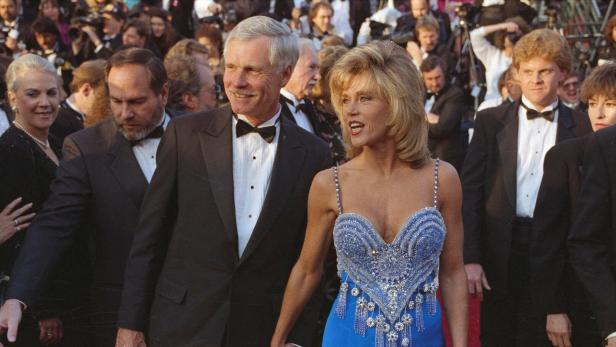 Jane Fonda und Ted Turner,, er im Smoking, sie in blauem Kleid, hält Händchen auf einem roten Teppich vor einer Menschenmenge.