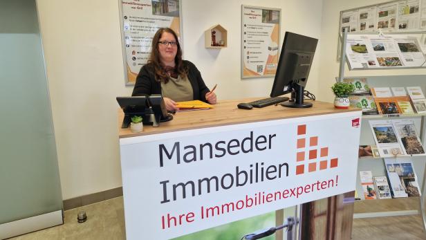 Eine Frau steht hinter einem Schild mit "Manseder Immobilien" 