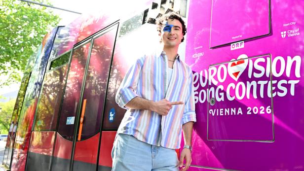 Ein junger Mann mit blauem Farbakzent im Gesicht posiert vor einer Straßenbahn mit "Eurovision Song Contest Vienna 2026"-Aufschrift.