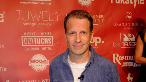 Oliver Pocher in Cordkleidung steht vor einer roten Wand mit Logos von Sponsoren.