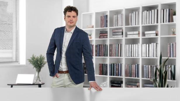 Mann im Anzug steht in modernem Büro mit Bücherregal und Pflanzen im Hintergrund.