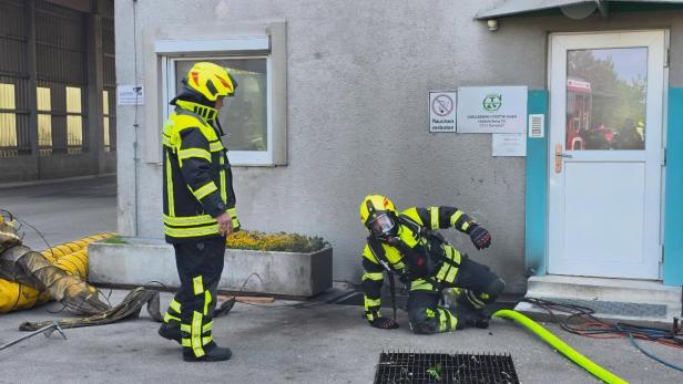 Zwei Feuerwehrleute in Schutzkleidung, einer steht, der andere kniet am Boden vor einem Gebäude mit Schläuchen und Ausrüstung.