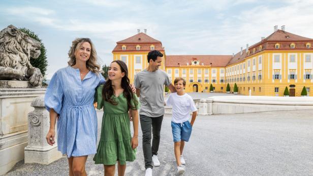 Vier Personen spazieren lachend vor dem gelben Schloss Hof und einer steinernen Löwenskulptur.