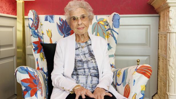 Lucy Waldstein mit Brille sitzt in einem bunten Sessel und lächelt freundlich.