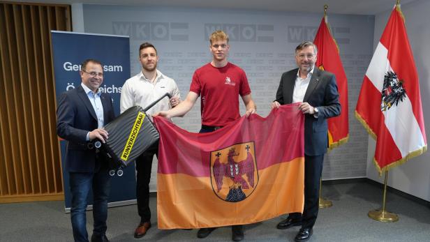Vier Männer stehen zusammen, halten eine rot-gelbe Fahne mit Adlerwappen und einen schwarzen Koffer mit gelbem Schriftzug.