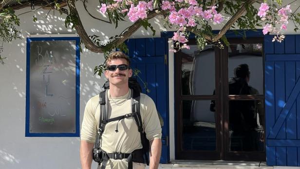 Ein junger Mann steht unter einem blühenden Baum, mit Rucksack, kurzer Hose und Sonnenbrille.