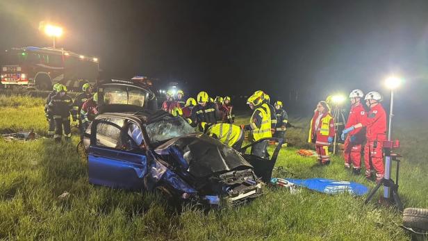 Mehrere Einsatzkräfte stehen nachts um ein stark beschädigtes Auto auf einer Wiese, Rettungsfahrzeuge und Scheinwerfer im Hintergrund.