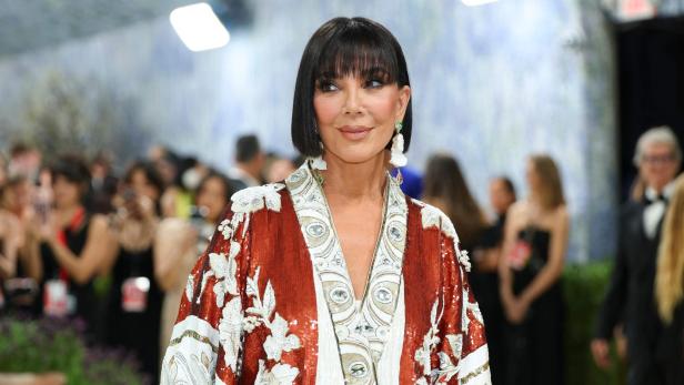 Kris Jenner trägt eine verzierte, rot-weiße Robe und blickt in einer Menschenmenge zur Seite.