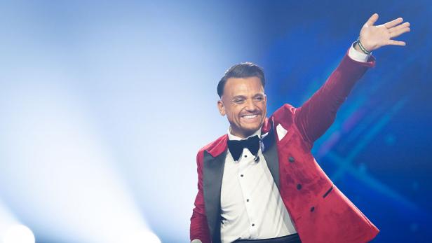 Menowin Fröhlich steht im roten Samtjackett mit Mikrofon auf der DSDS-Bühne und winkt lächelnd ins Publikum.