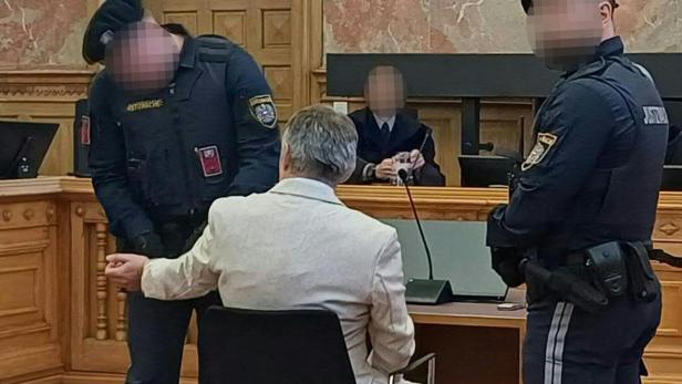 Ein Mann in einem hellen Anzug sitzt im Gerichtssaal, bewacht von zwei Justizwachebeamten.
