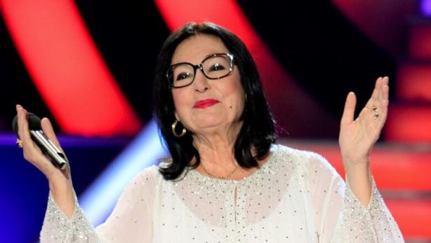 Nana Mouskouri mit Brille und weißer Bluse hält ein Mikrofon in der Hand.