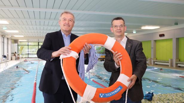 Zwei Männer im Anzug halten einen Rettungsring am Beckenrand eines Hallenbads, im Hintergrund schwimmen Menschen.
