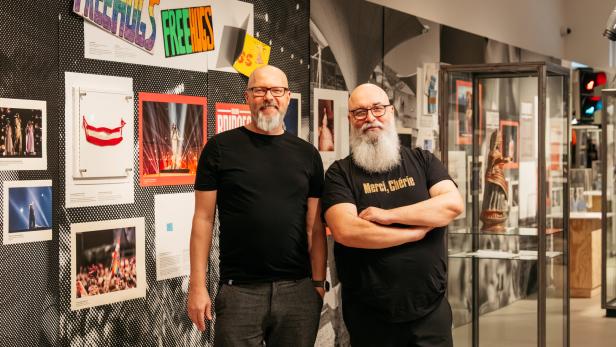 Marco Schreuder und Alkis Vlassakakis mit Brille und Bart stehen lächelnd in einer Ausstellung mit Fotos, Schildern und Vitrinen.