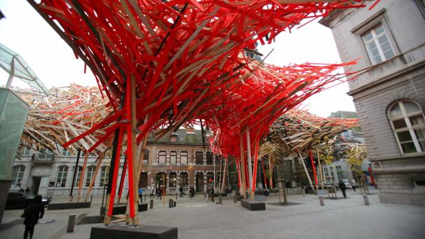 Eine Installation aus roten und hellen Holzstreifen überspannt einen Platz in einer Stadt.