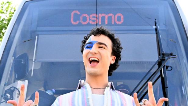 Eine Person mit blauem Gesichtsmakeup und bunter Kleidung steht lachend vor einer Straßenbahn mit der Anzeige „Cosmo“.