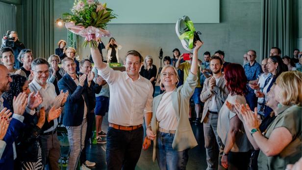 Zwei Personen stehen jubelnd inmitten applaudierender Menschen, eine hält Blumen, die andere ein Paar Sportschuhe hoch.