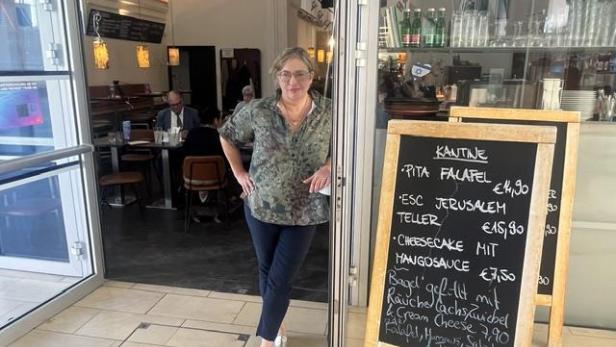 Frau steht am Eingang eines Cafés neben einer Tafel mit Speisekarte, Gäste sitzen im Hintergrund.