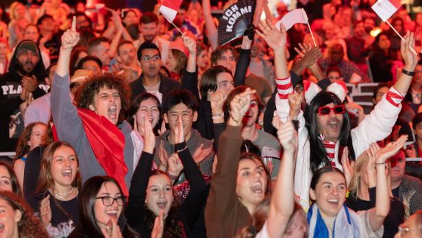 Jubelnde Menschenmenge mit rot-weißen Fahnen, viele lachen, klatschen und feiern ausgelassen.