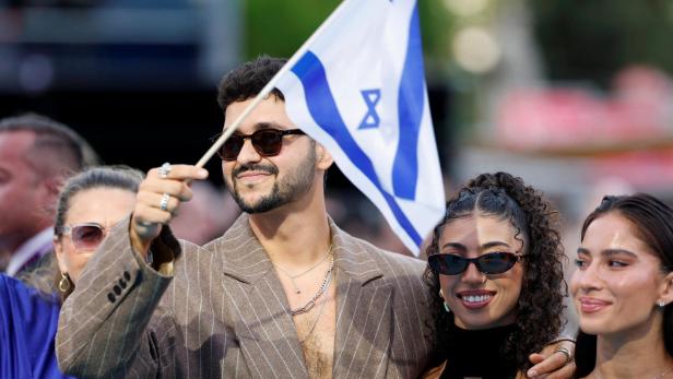Ein Mann mit Sonnenbrille hält eine israelische Flagge, neben ihm stehen zwei lächelnde Frauen im Freien.